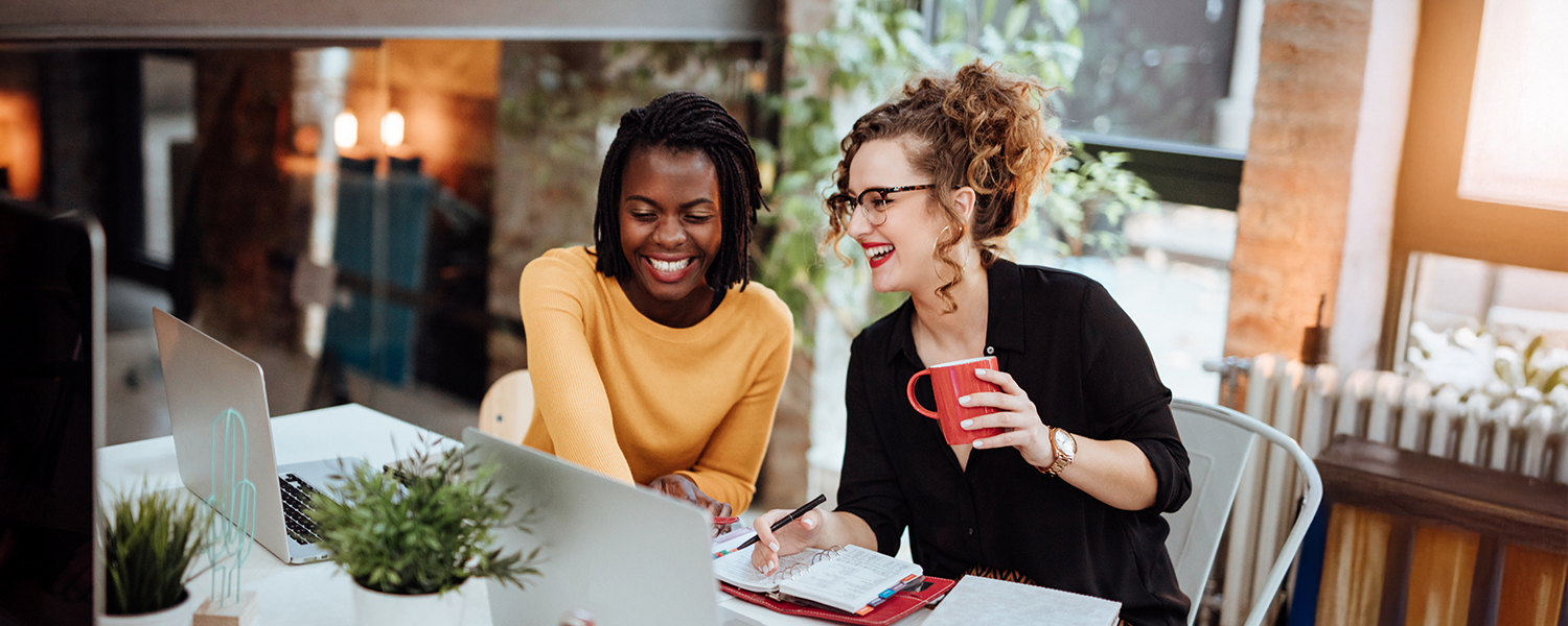 Photo of two women working on laptops, they are smiling and collaborating