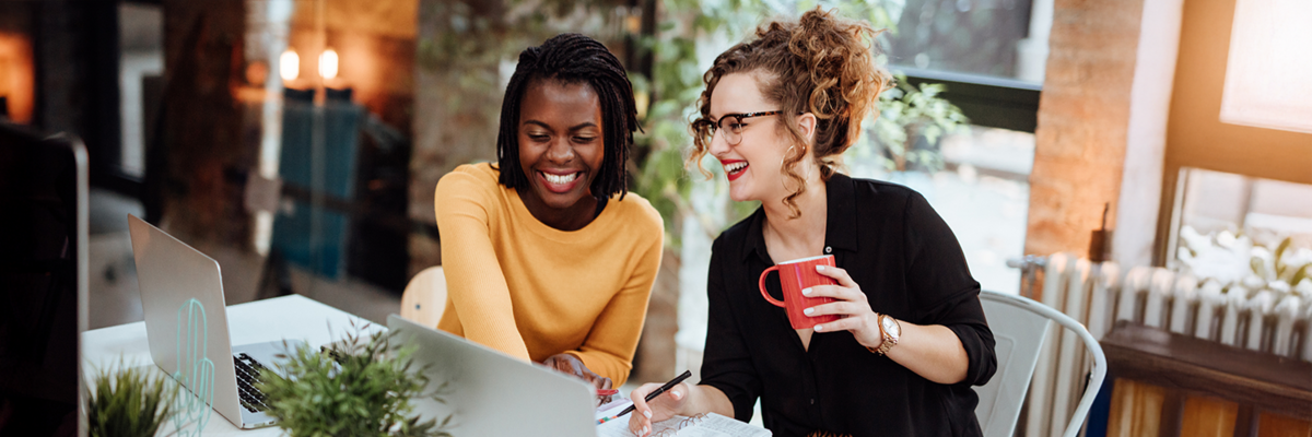 Photo of two women working on laptops, they are smiling and collaborating