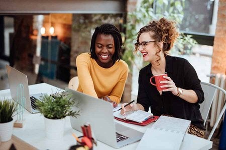 Photo of two women working on laptops, they are smiling and collaborating
