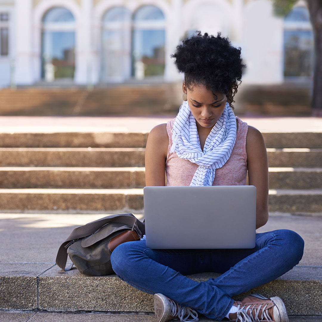 Image of a college student sitting on the ground at their university while on their computer